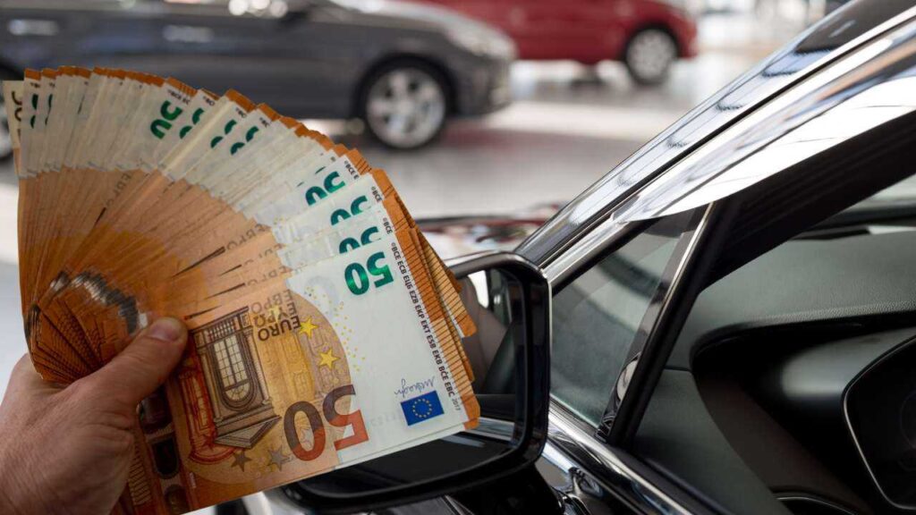 persona che tiene banconote da 50€ in primo piano e auto sullo sfondo