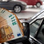 persona che tiene banconote da 50€ in primo piano e auto sullo sfondo