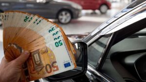 persona che tiene banconote da 50€ in primo piano e auto sullo sfondo