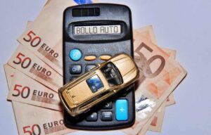 Una macchina giocattolo sopra a una calcolatrice con critto bollo auto, a sua volta sopra a banconote da 50€